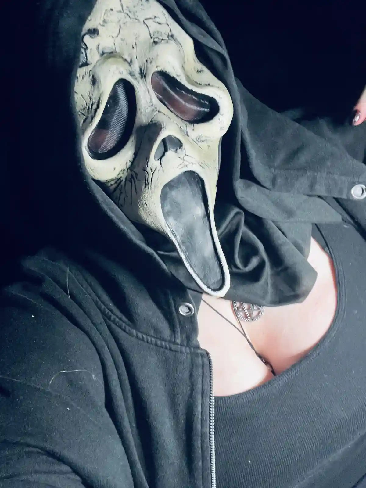 A person wearing a black hoodie and a black zip-up jacket has a white, cracked, screaming Ghostface mask from the Scream…