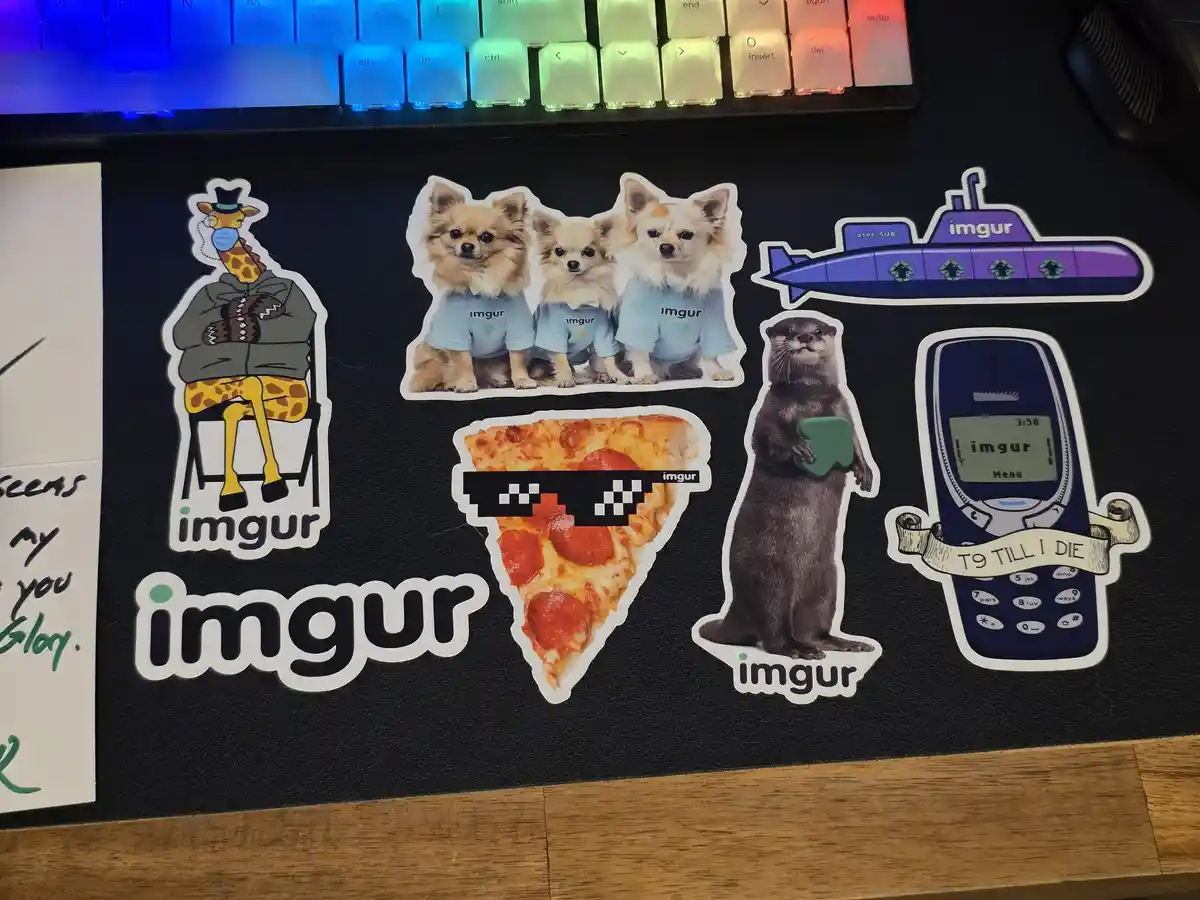 A collection of Imgur-branded stickers on a black mousepad, featuring a giraffe in a hoodie, three chihuahuas in blue…