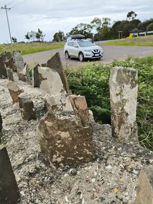 A silver SUV with a roof box drives along a paved road, viewed from behind a row of weathered, broken stone posts. The…