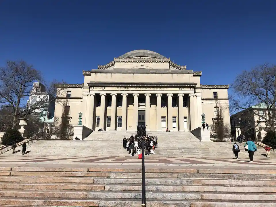 A large group of students is posing for a photo on the wide stone steps in front of the grand, neoclassical Columbia…