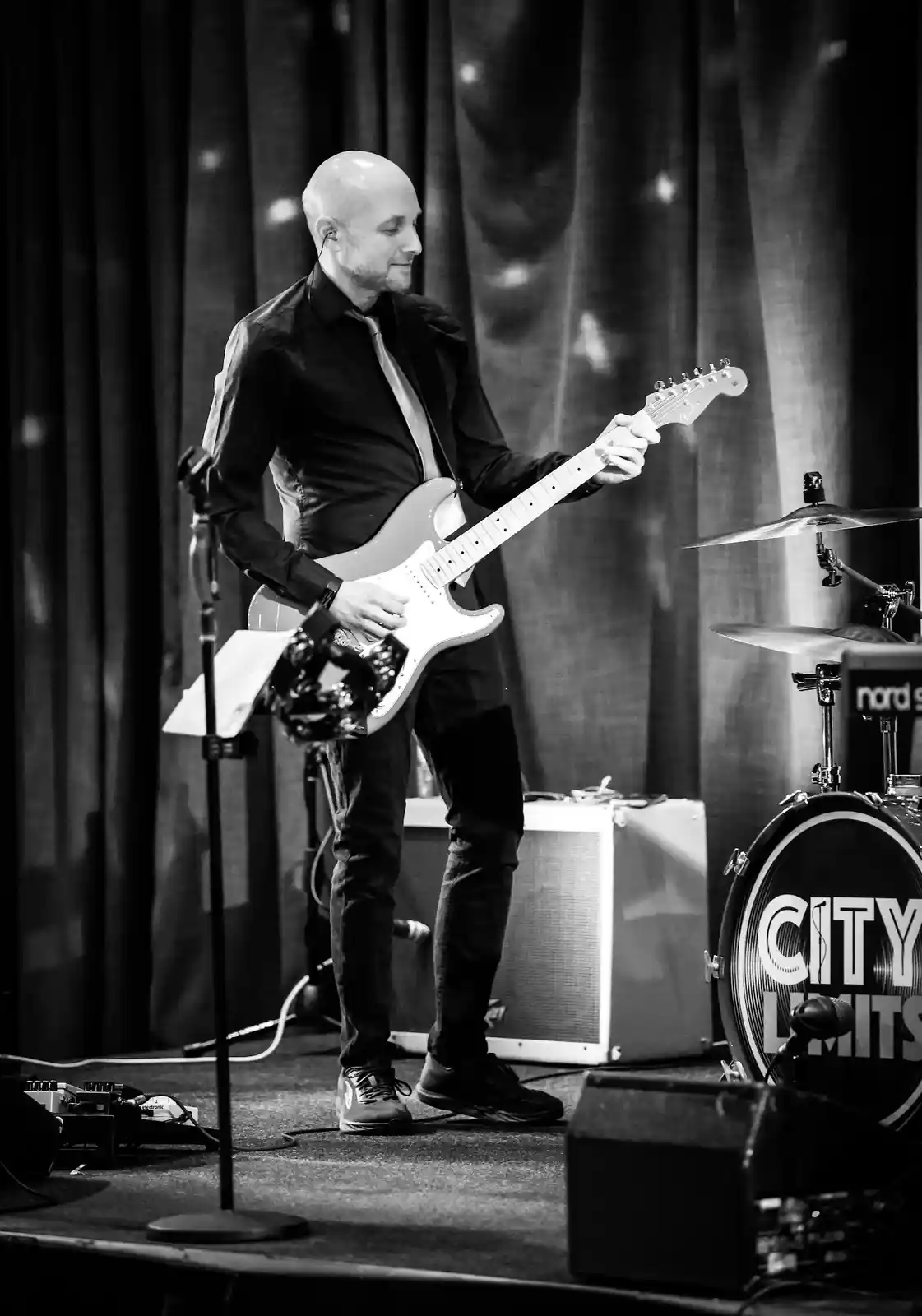 A black and white photo of a bald man in a black shirt and tie playing a white electric guitar on stage, with a drum kit…