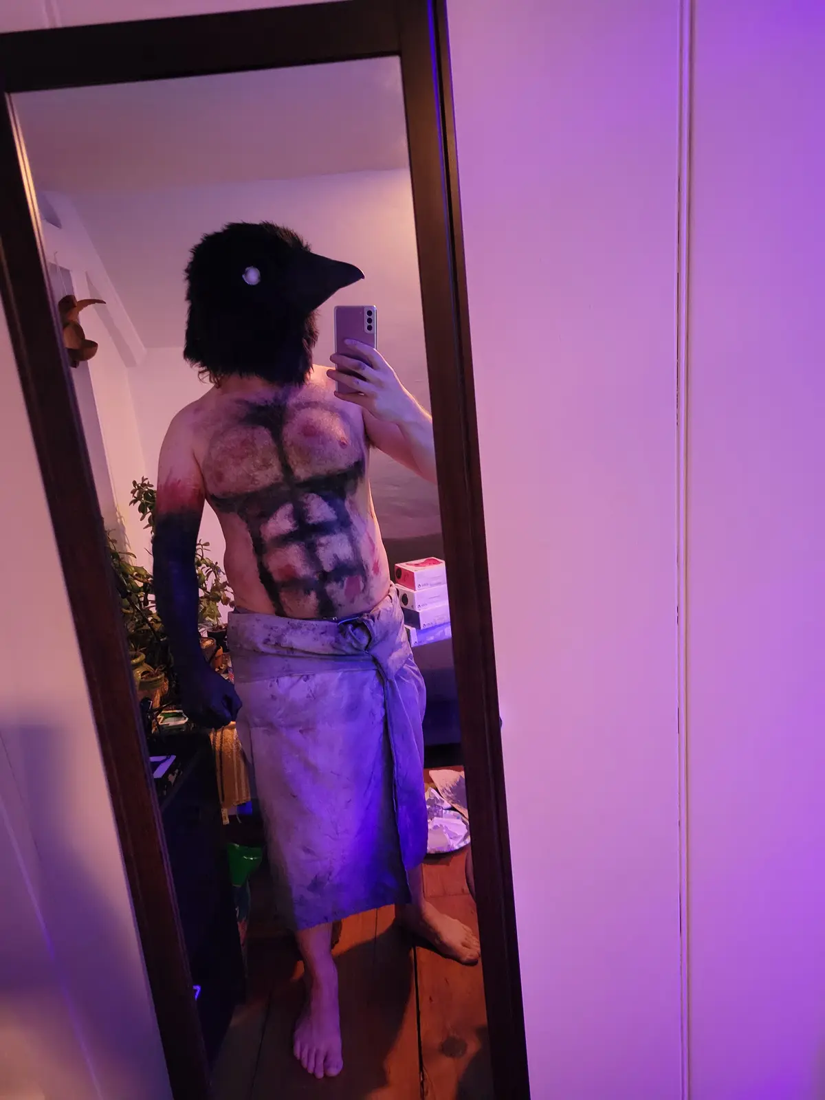 A person wearing a large black crow mask and a towel around their waist takes a mirror selfie, their bare chest painted with…