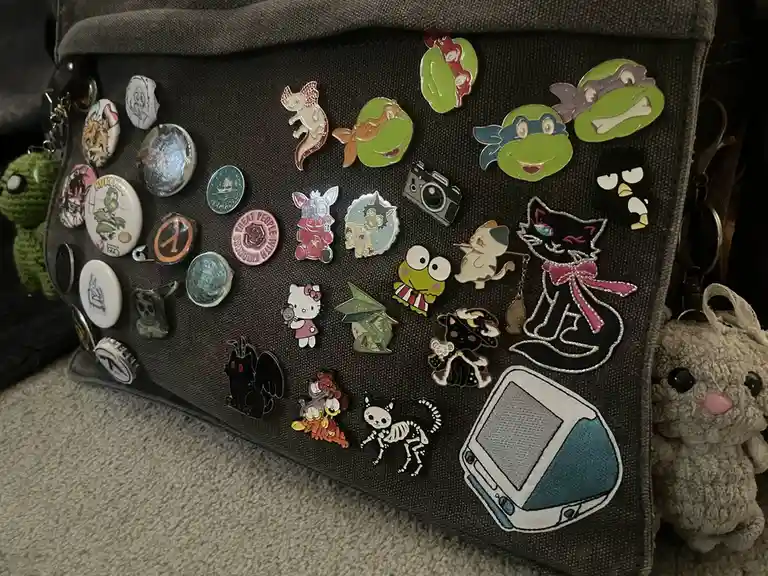 A dark gray fabric bag or pouch is densely covered with dozens of colorful enamel pins featuring various characters and…