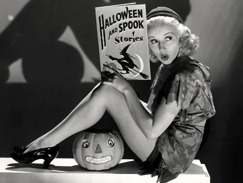 A vintage black-and-white photo shows a woman in a Halloween costume sitting on a platform with a carved jack-o'-lantern…