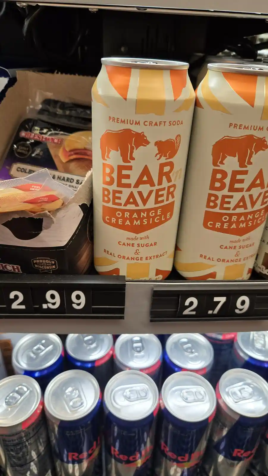 Two cans of Bear'n Beaver Orange Creamsicle soda sit on a store shelf with price tags showing $2.99 and $2.79. Below them…