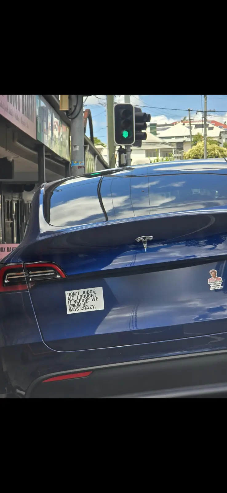 The image shows the rear of a blue Tesla vehicle parked at a traffic light. A white rectangular sticker on the trunk reads…