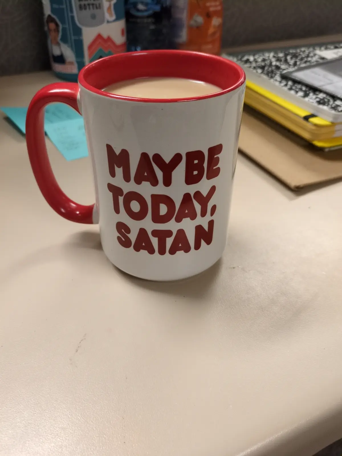 A white mug with a red rim and handle sits on a desk, filled with a light brown beverage. The mug has bold red text reading…