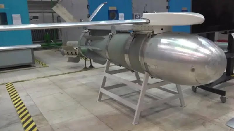 a large, silver, cylindrical missile with wings and tail fins is displayed on a metal stand inside a factory or hangar. The…