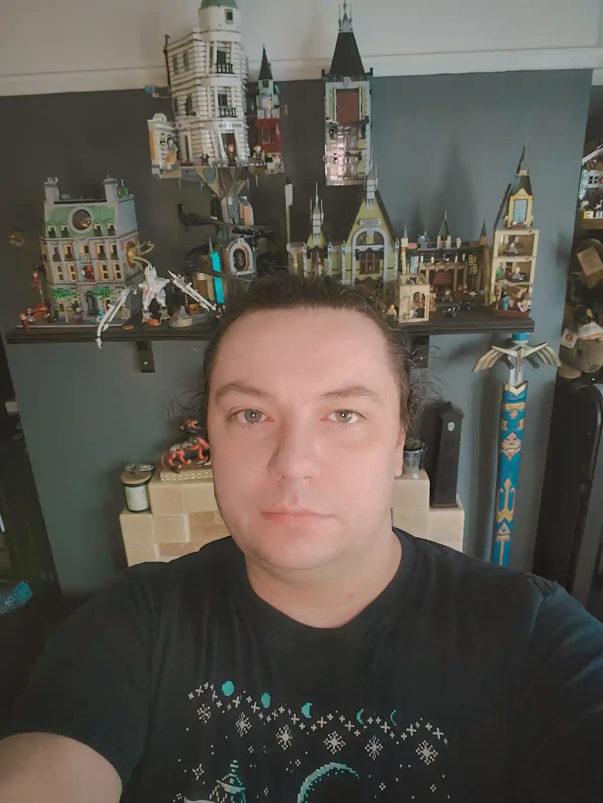 A man with a neutral expression takes a selfie in front of a large, cluttered shelf filled with elaborate LEGO castle and…