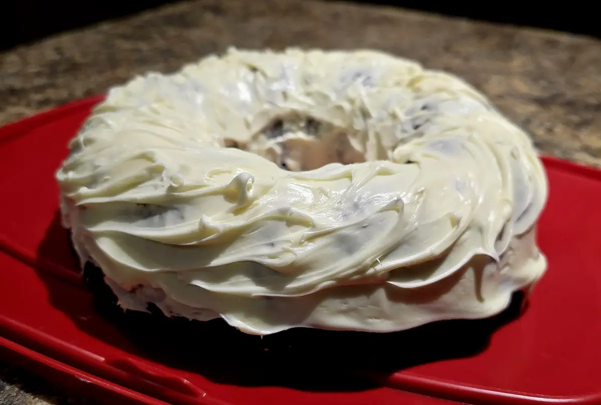 A close-up photo shows a ring-shaped bagel heavily frosted with creamy white cream cheese, sitting on a bright red plastic…