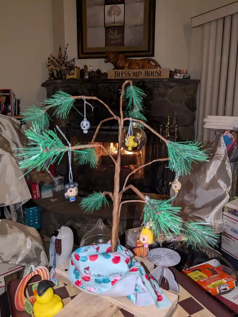 A small, artificial Christmas tree with green pine needles and various ornaments hangs in front of a lit stone fireplace…