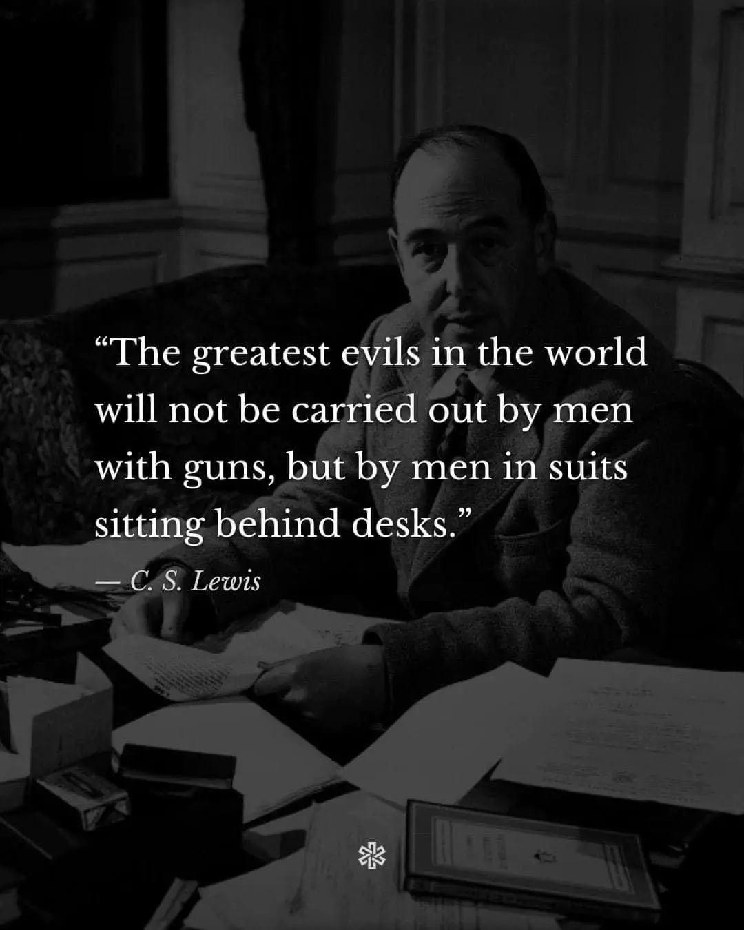 A black-and-white photo shows C. S. Lewis seated at a cluttered desk, surrounded by papers and books, looking directly at…