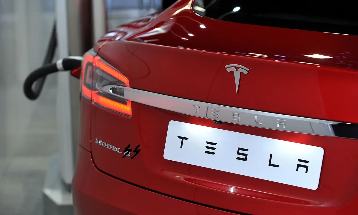 A close-up of the rear of a red Tesla Model 3, plugged into a charging station, with the official Tesla logo and 'MODEL 3'…
