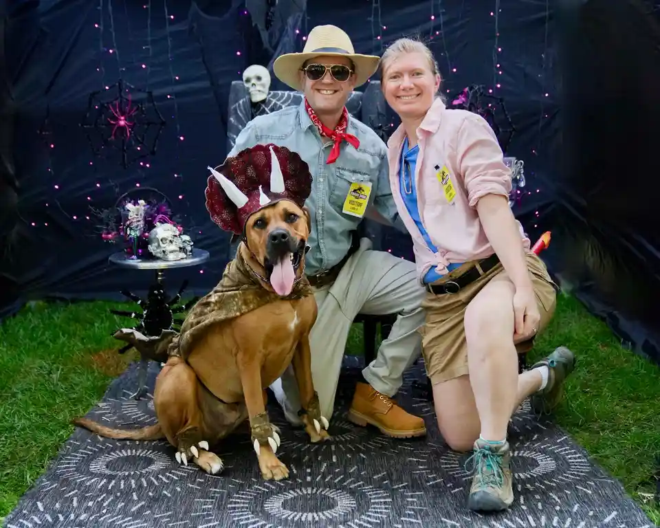 A brown dog wearing a triceratops costume with horns and claws sits between a man and a woman dressed as Jurassic Park…