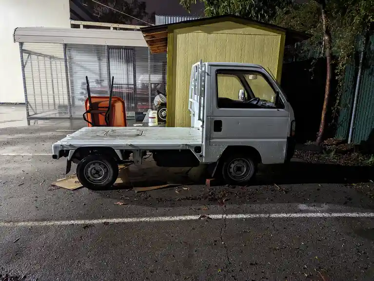 A small white flatbed truck is parked at night in a parking lot, partially blocking a yellow shed. The truck's rear wheels…