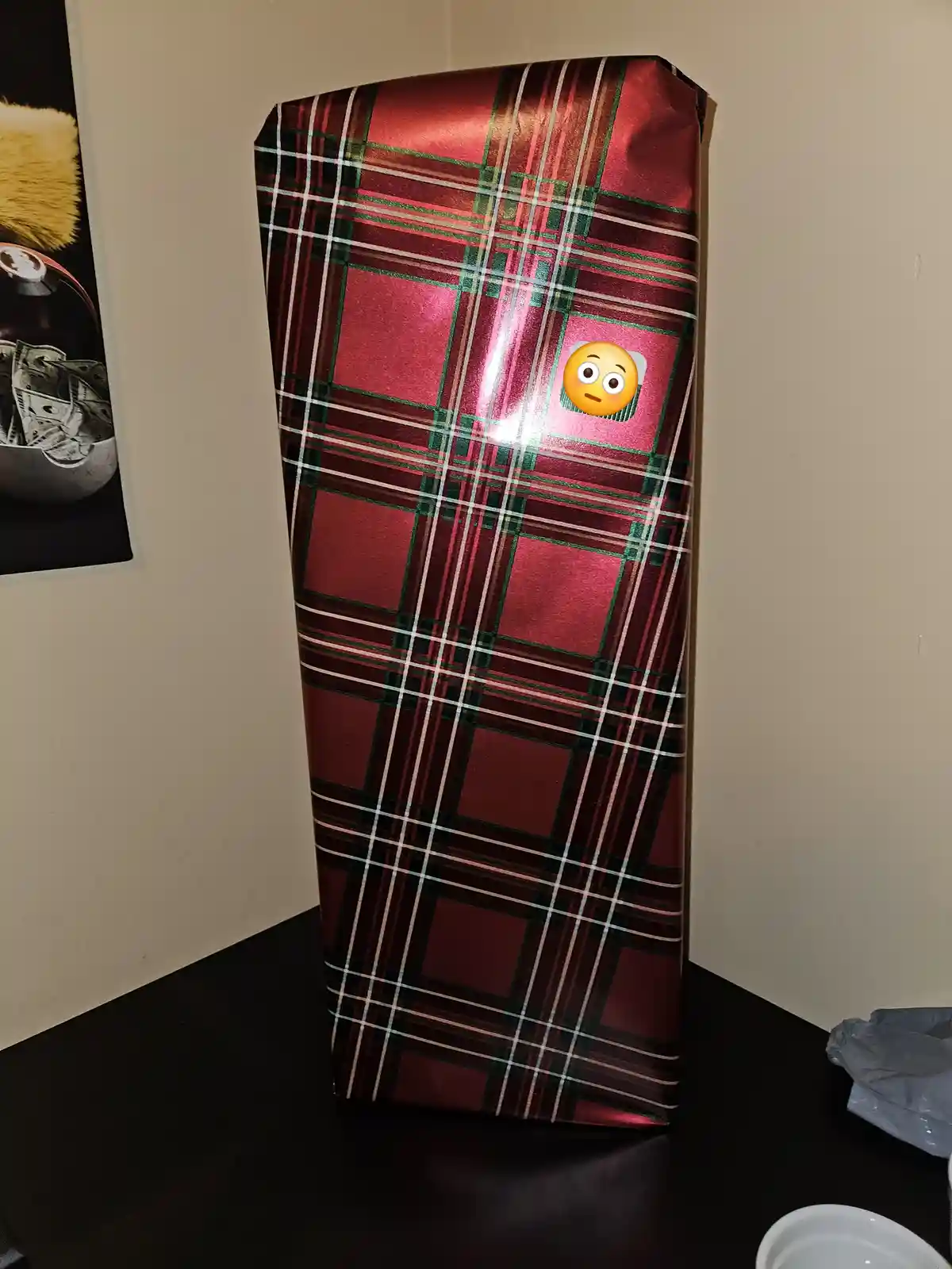 A tall, rectangular gift wrapped in shiny red, green, and white plaid paper stands on a dark table. A surprised face emoji…