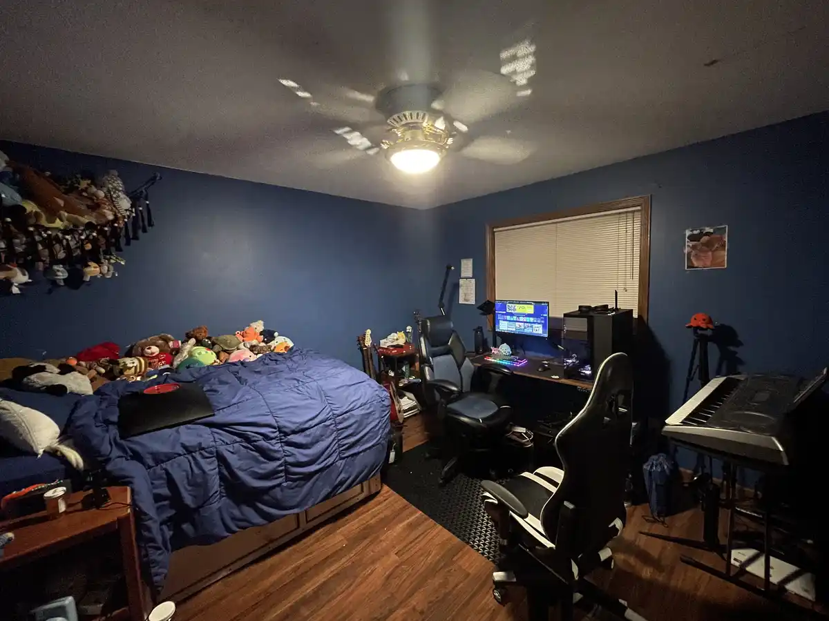 A dimly lit bedroom with blue walls, a bed covered in a blue comforter and stuffed animals, a gaming setup with two chairs…