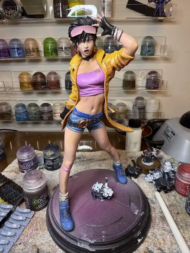A painted figurine of a female character in a yellow jacket, purple top, and denim shorts stands on a purple base surrounded…