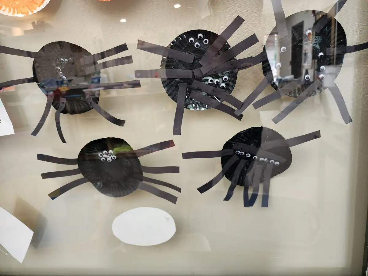 Four handmade black paper plate spiders with googly eyes and black paper legs are taped to a glass surface, likely a…