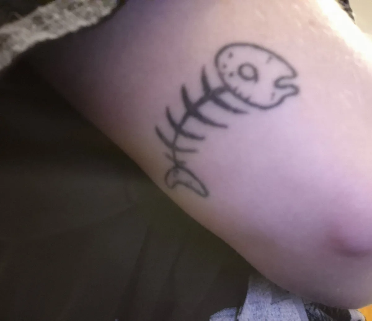 A close-up photo shows a simple black ink tattoo on human skin, depicting a cartoon-style fish skeleton with a round head…