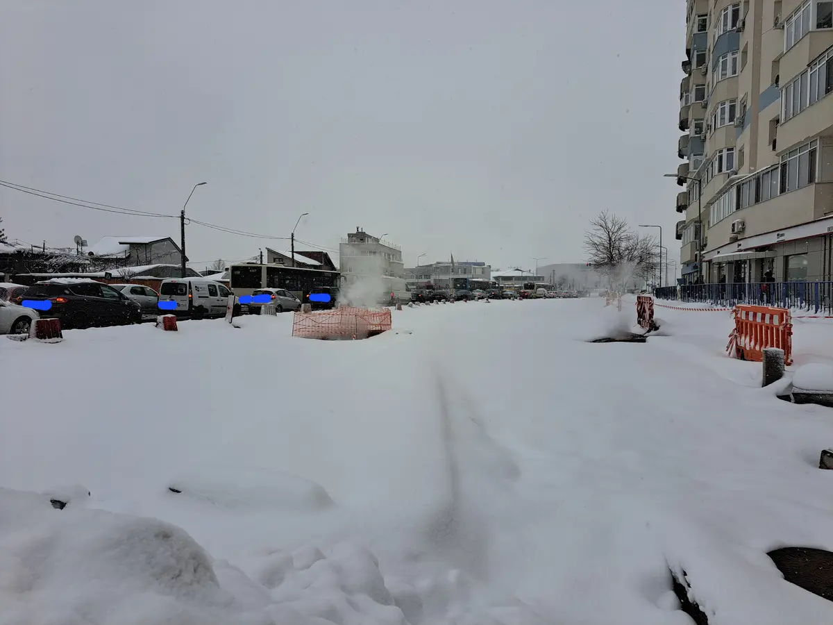 A snowy urban street with cars and buses partially buried under deep snow, orange construction barriers, and a visible path…
