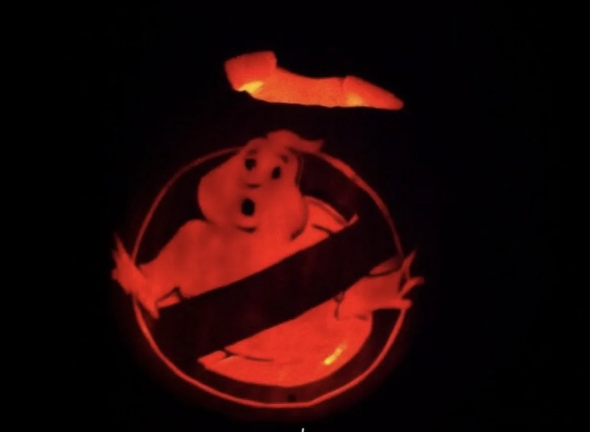 A carved pumpkin illuminated from within, displaying the Ghostbusters logo with a cartoon ghost trapped inside a circular…