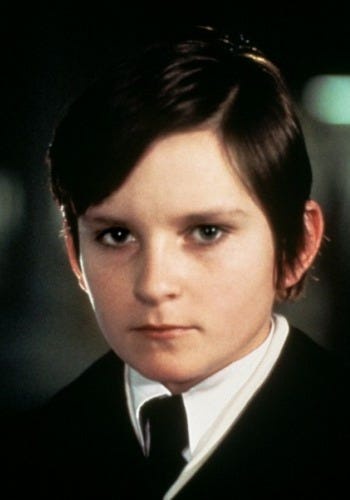 A still of a young boy with dark hair and a serious expression, wearing a black suit and white collared shirt. The overlaid…