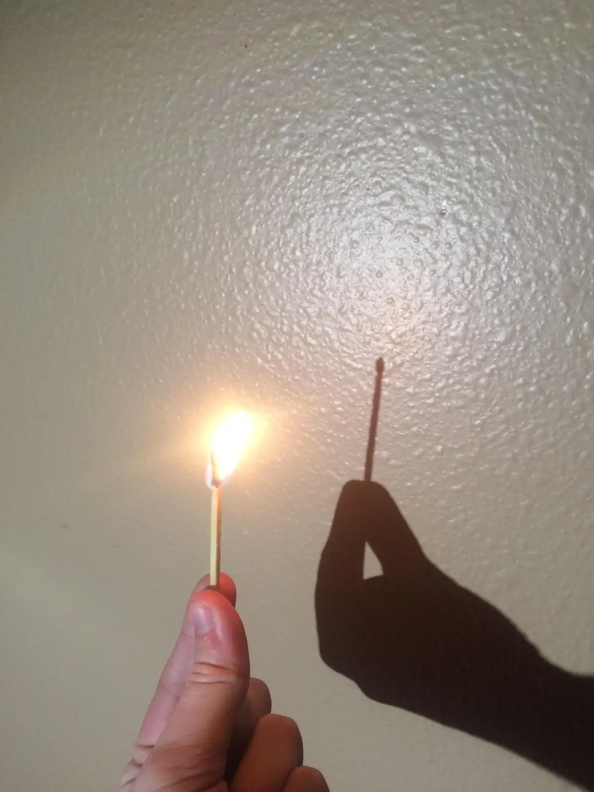 A hand holds a lit matchstick against a textured white wall, casting a distinct shadow that appears as a simple stick shape…