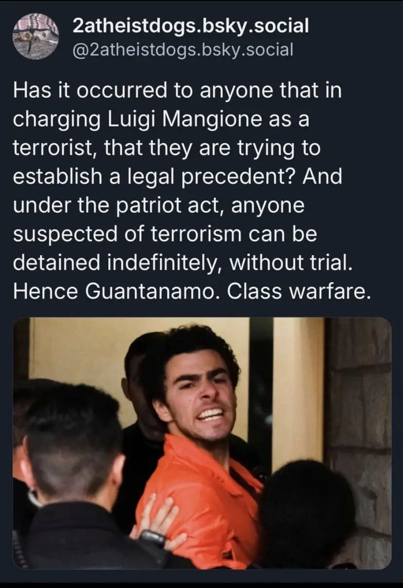 A man in an orange jumpsuit is being escorted by several people in dark clothing, his face contorted in an expression of…