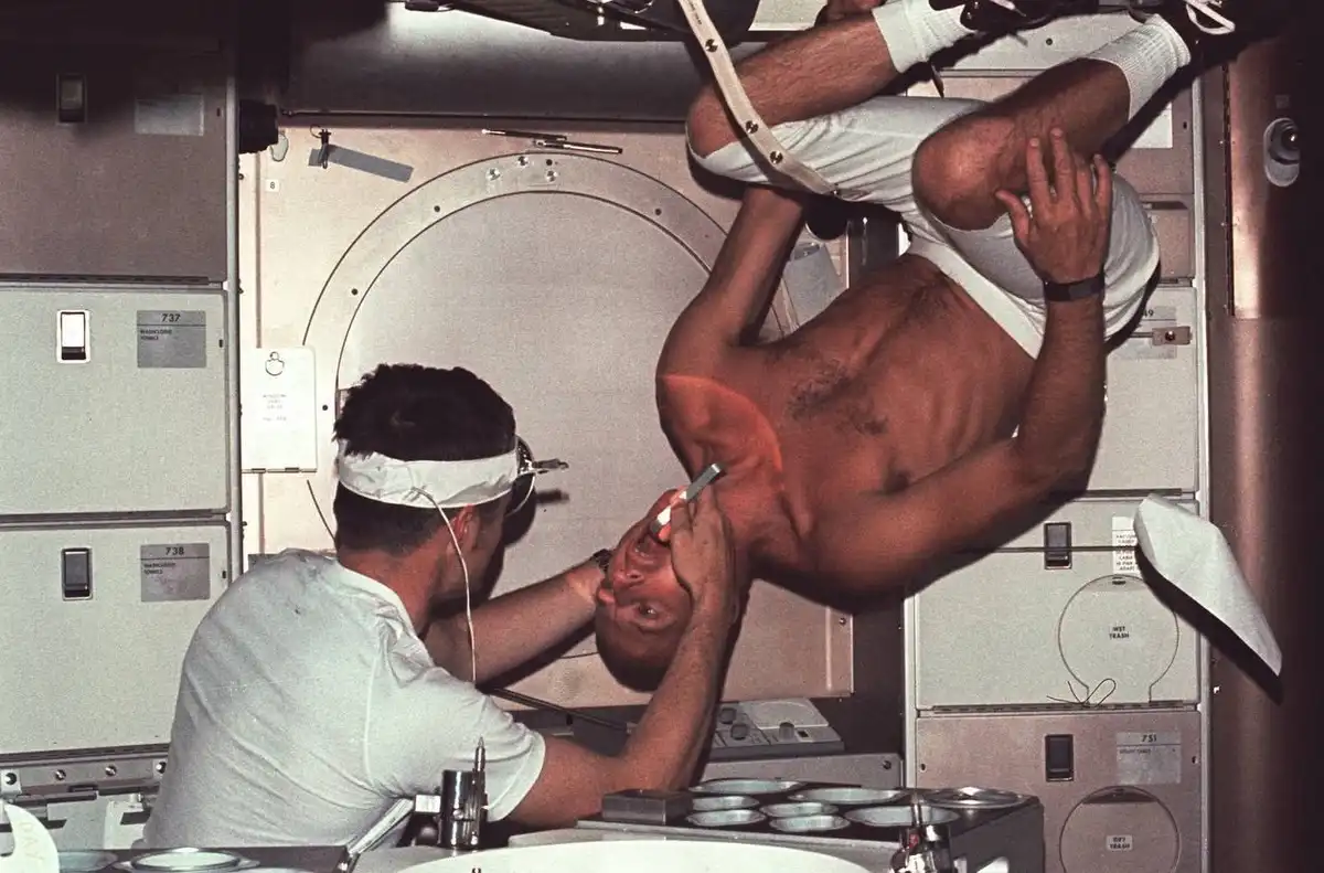 A shirtless man is suspended upside down in a spacecraft-like environment while another man in a white shirt and headband…