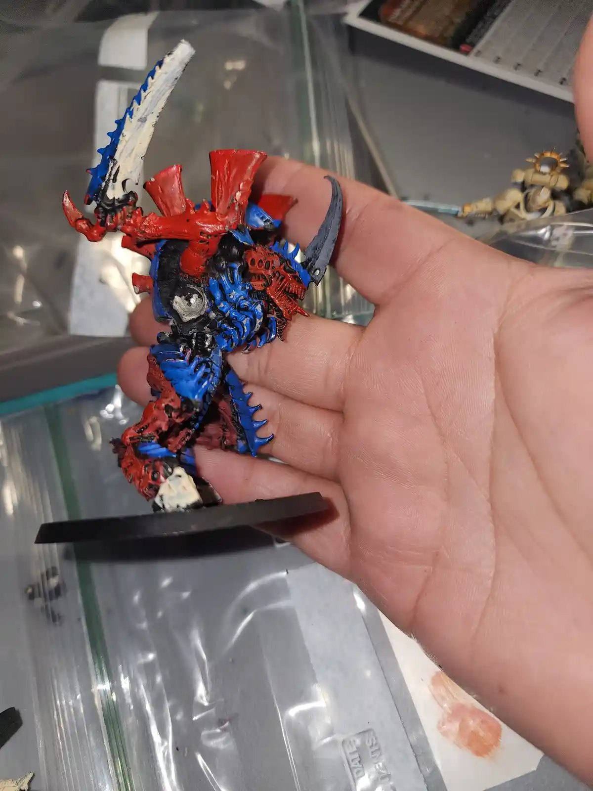 A hand holds a roughly painted miniature figurine, predominantly red and blue with black and white accents, showing uneven…