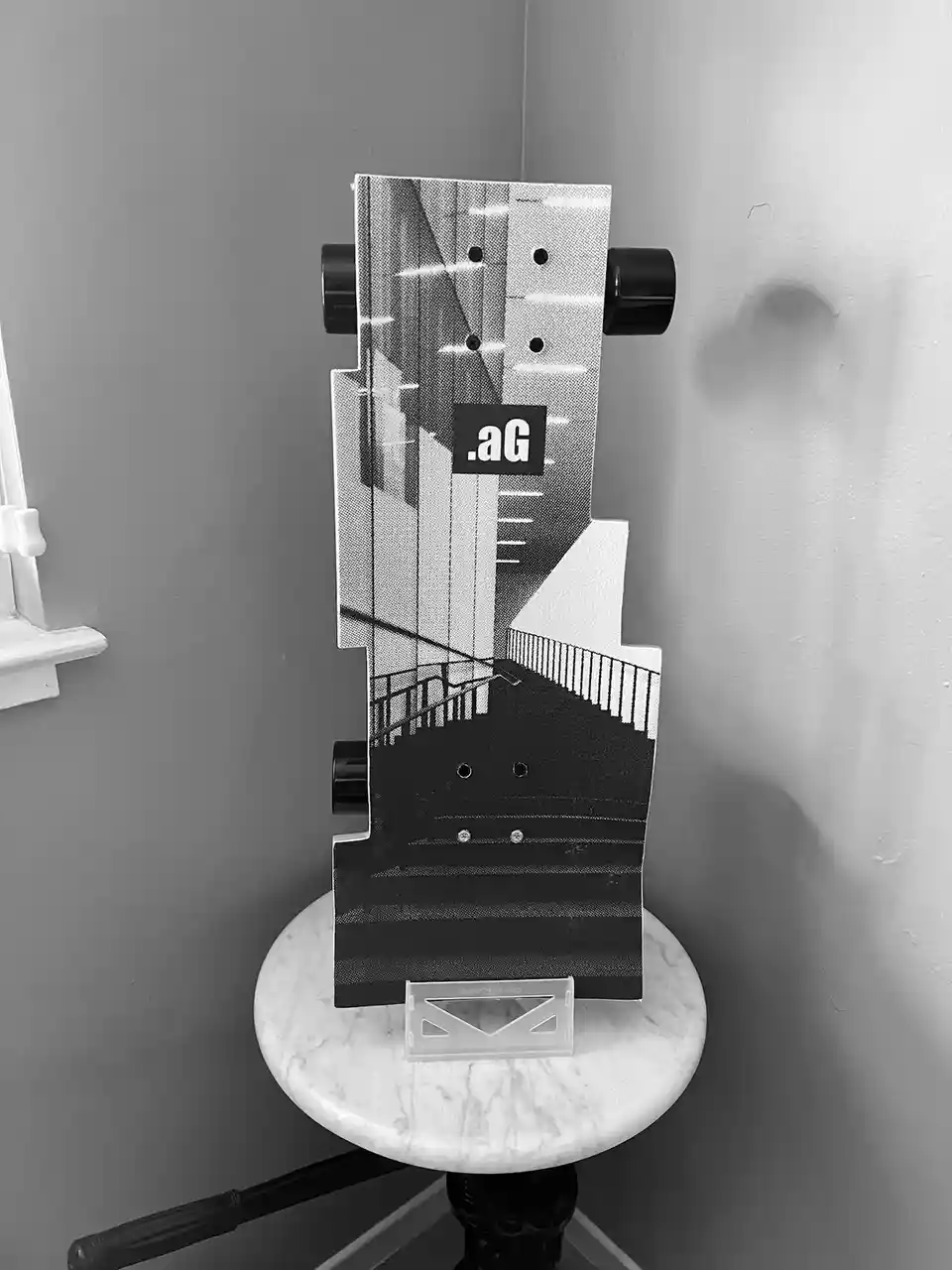 A black and white skateboard deck with a graphic of a staircase and cityscape is standing upright on a small white marble…