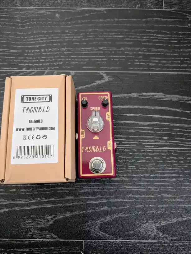 A red Tone City Tremble guitar effects pedal sits next to its cardboard packaging on a dark wood-grain surface. The pedal…