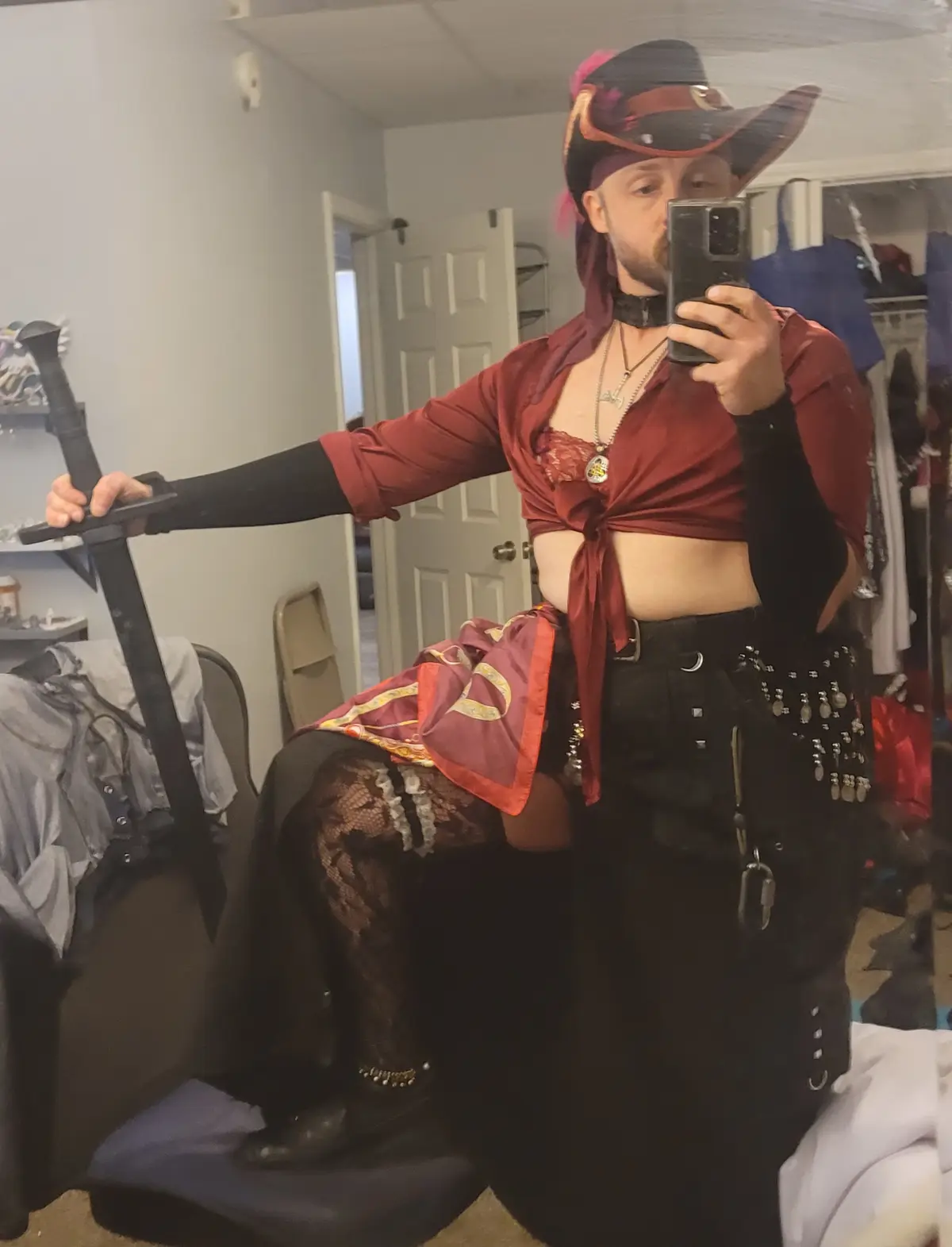 A person in a room takes a mirror selfie while dressed in a pirate costume, holding a sword. They wear a red crop top tied…