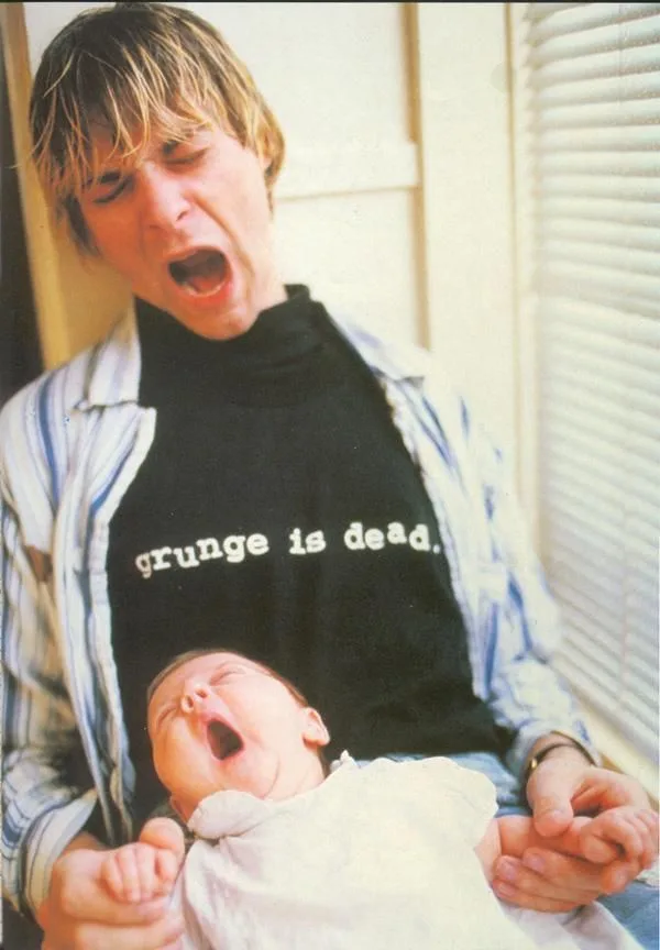 A man with messy blonde hair wearing a black t-shirt that reads 'grunge is dead.' is holding a baby. Both the man and the…