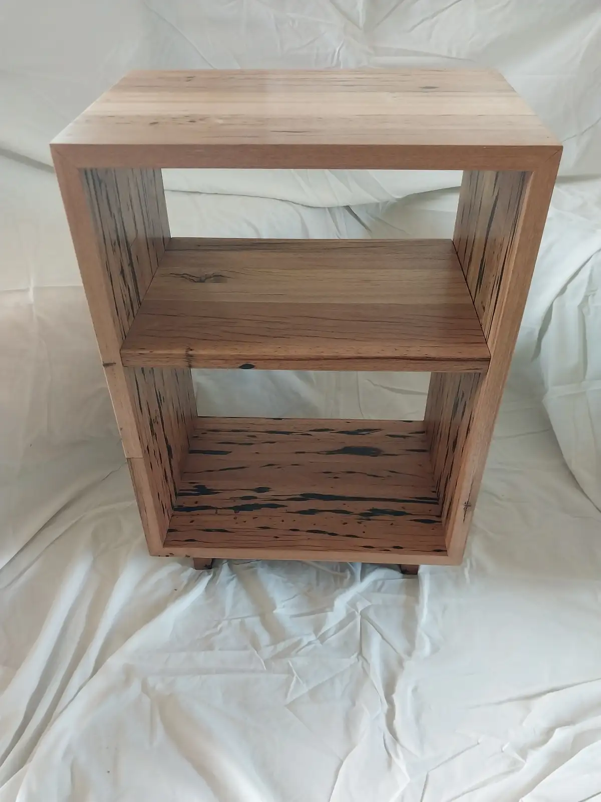 A wooden cube-shaped shelf with three levels sits on a white fabric backdrop. The top two shelves have a natural light wood…