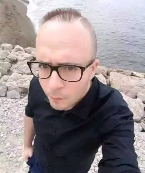 A man with a very short buzz cut and a single strip of hair standing on a rocky shoreline, looking directly at the camera…