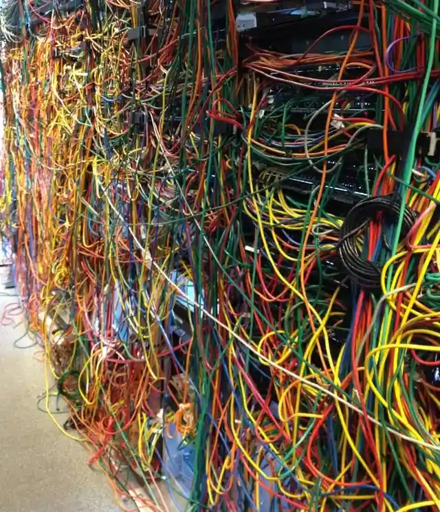 A wall of server racks is completely overwhelmed by a chaotic tangle of multicolored cables—red, yellow, green, blue, and…