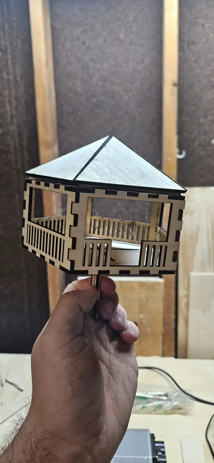 A hand holds up a small, intricately laser-cut wooden model of a gazebo or tiny house with a pointed roof and railings…