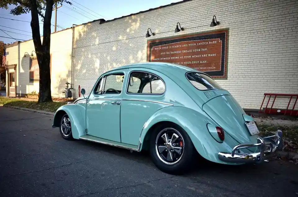 A mint green classic Volkswagen Beetle is parked on a street in front of a white brick building. On the building wall, a…