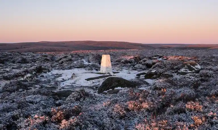 A serene, snow-dusted landscape at sunrise or sunset features a white, truncated conical trig point standing alone in a…