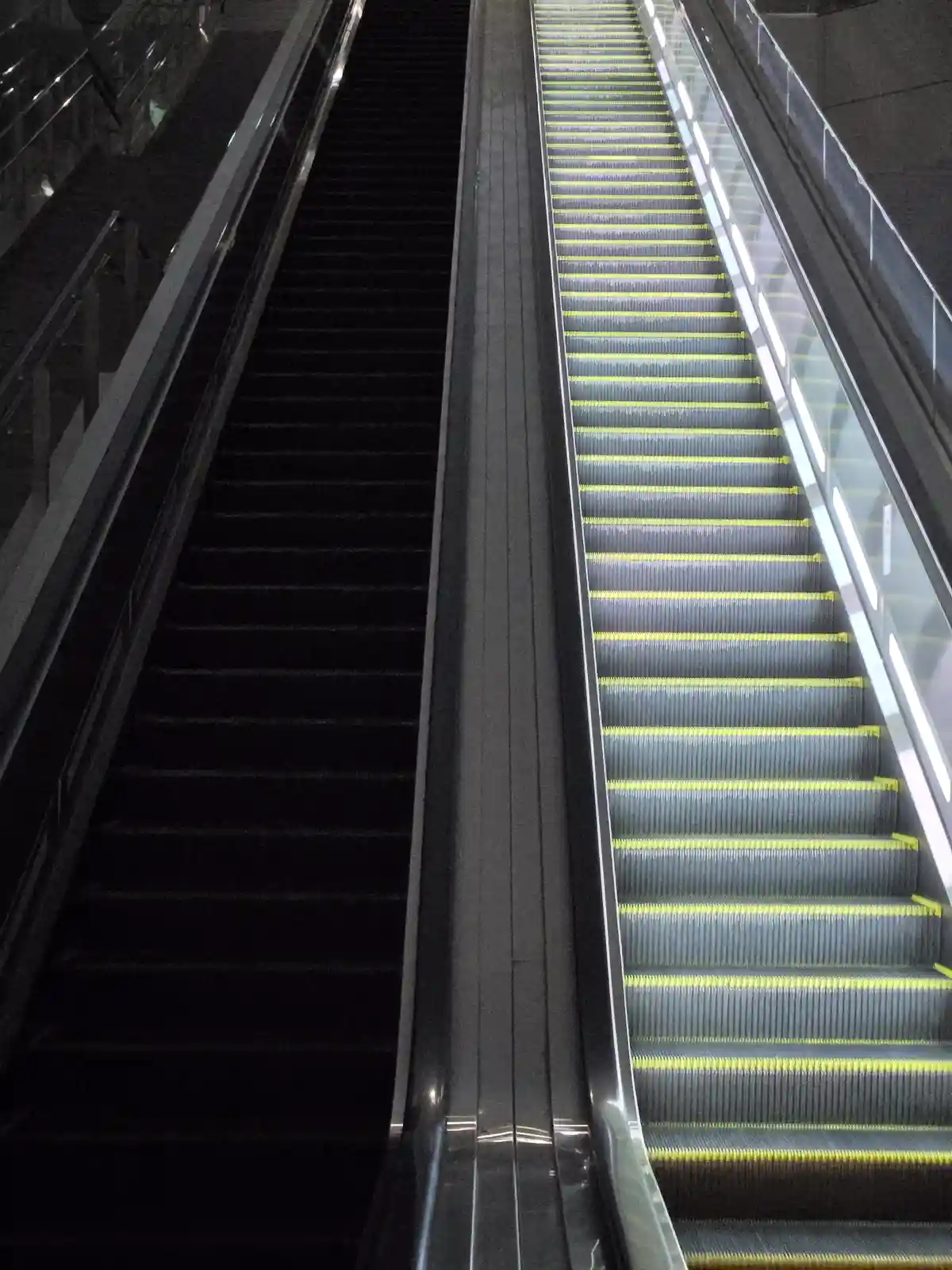 A side-by-side photo of two escalators in a public space, one moving upward with yellow safety stripes and the other moving…