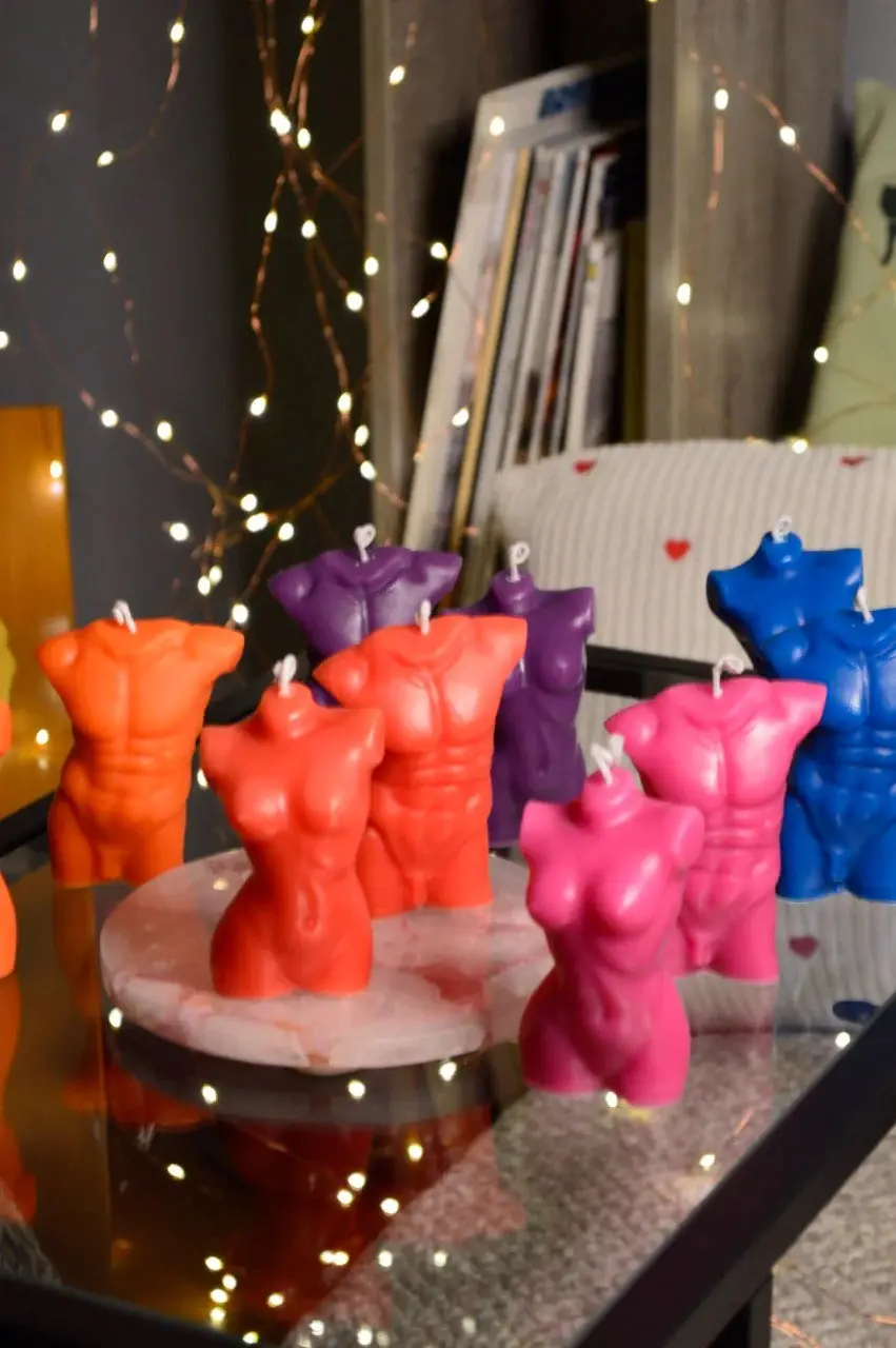 A collection of brightly colored candles shaped like muscular male torsos and curvaceous female torsos are arranged on a…