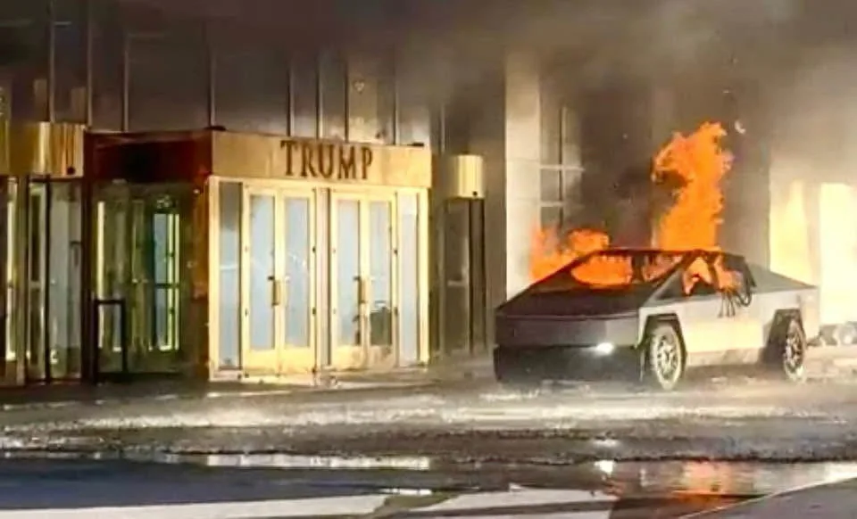 A Tesla Cybertruck is engulfed in flames on a wet street outside a building with a golden 'TRUMP' sign above its entrance…