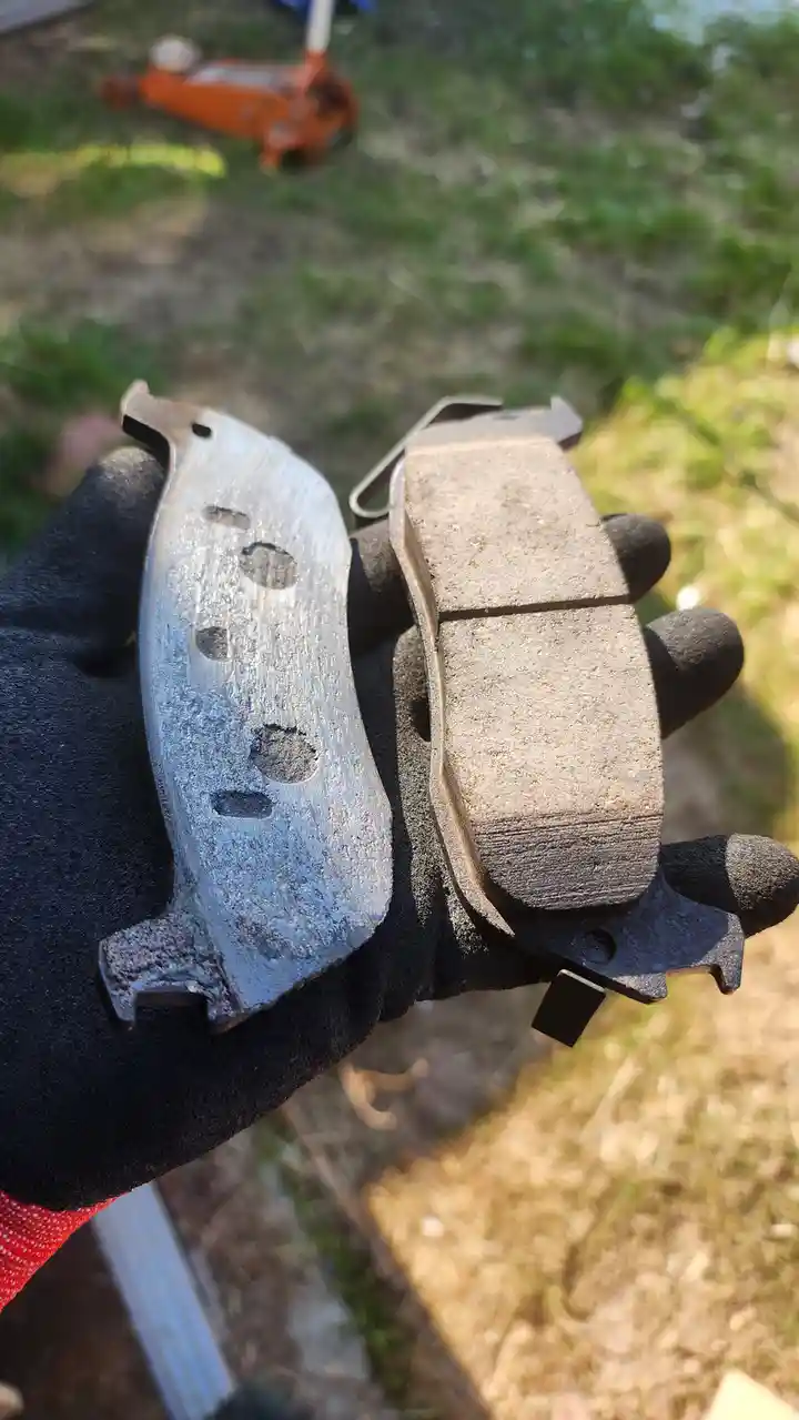 A gloved hand holds two brake pads side by side outdoors; one is heavily worn and corroded, the other is new and intact. The…