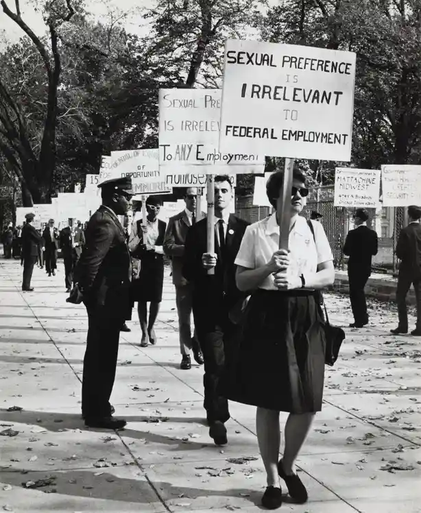 A black-and-white photograph shows a 1960s LGBTQ+ rights protest with demonstrators holding signs, including one prominently…