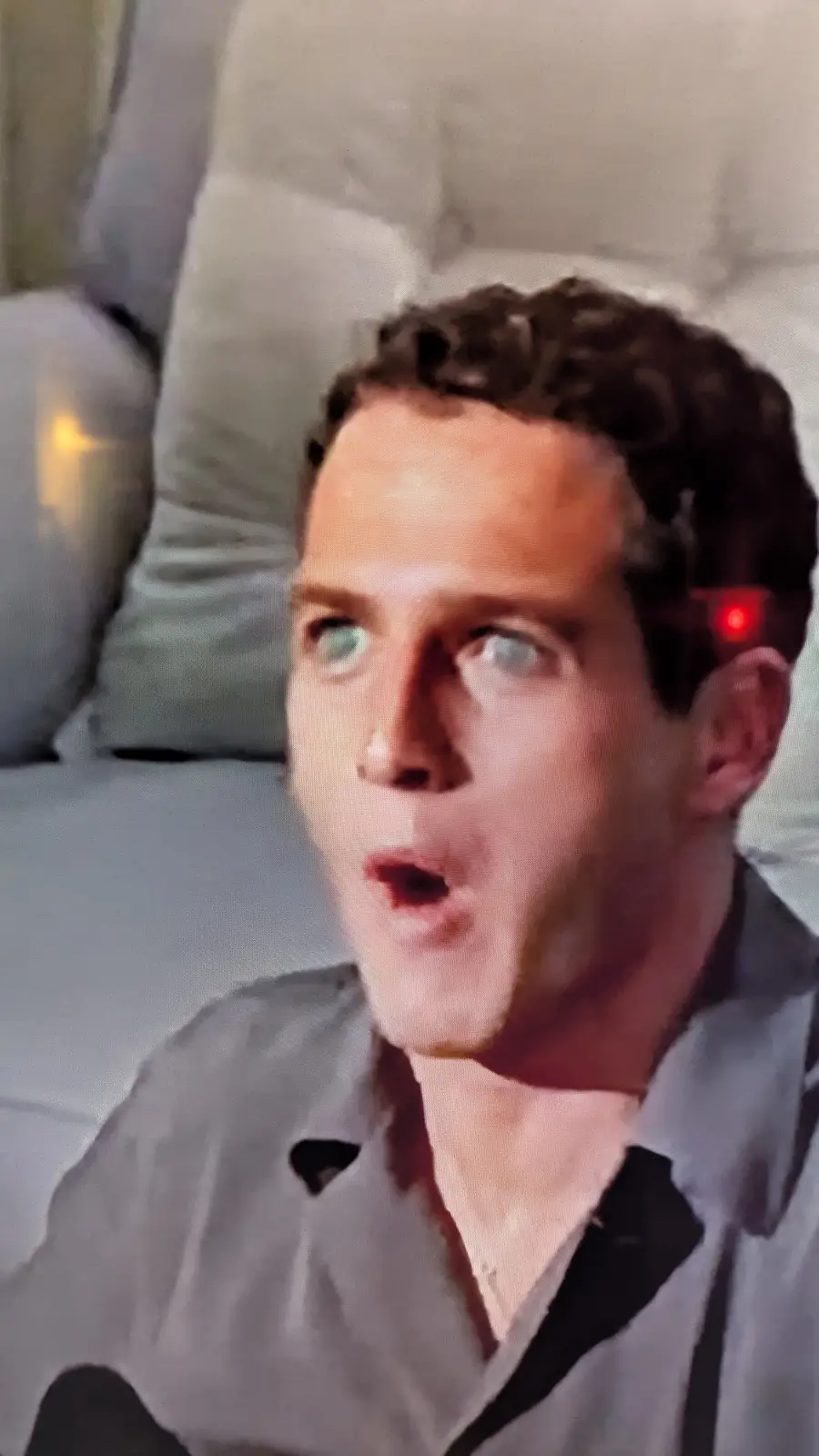 A man with wide eyes and an open mouth sits on a couch, looking shocked. A red light is digitally added to his temple. The…