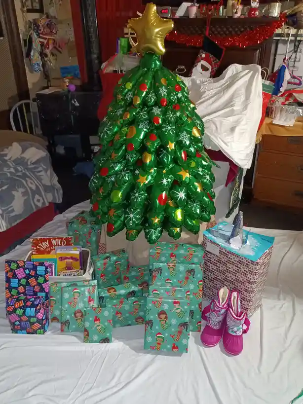 A large, green, inflatable Christmas tree made of balloons, topped with a gold star, stands on a bed surrounded by numerous…
