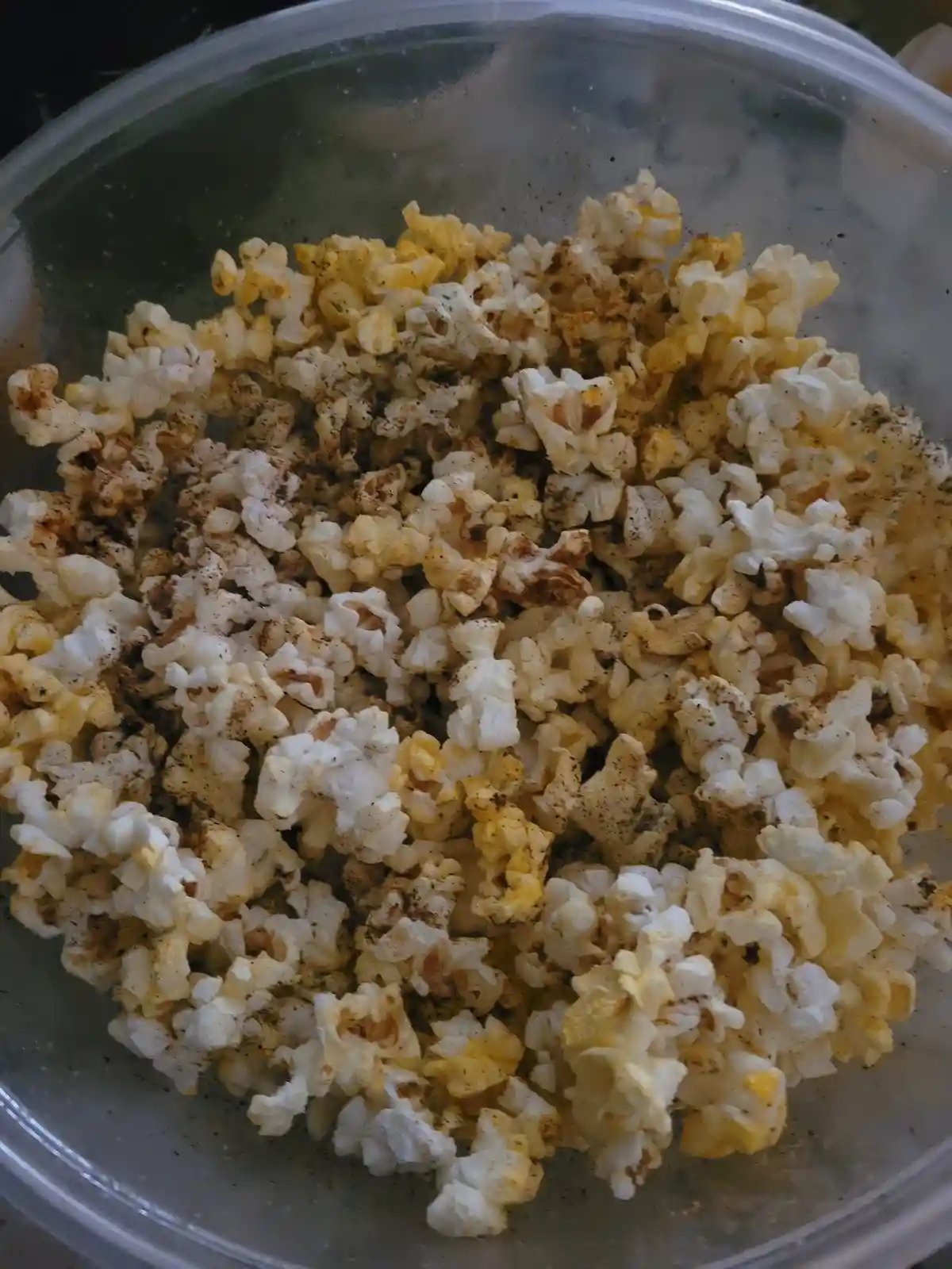 A close-up photo shows a clear plastic bowl filled with popcorn kernels that are heavily speckled with dark seasoning…