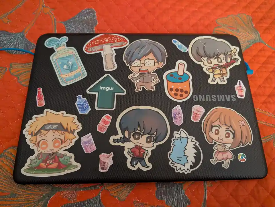 A black laptop covered in numerous colorful stickers, including anime characters like Naruto and a chibi version of a…